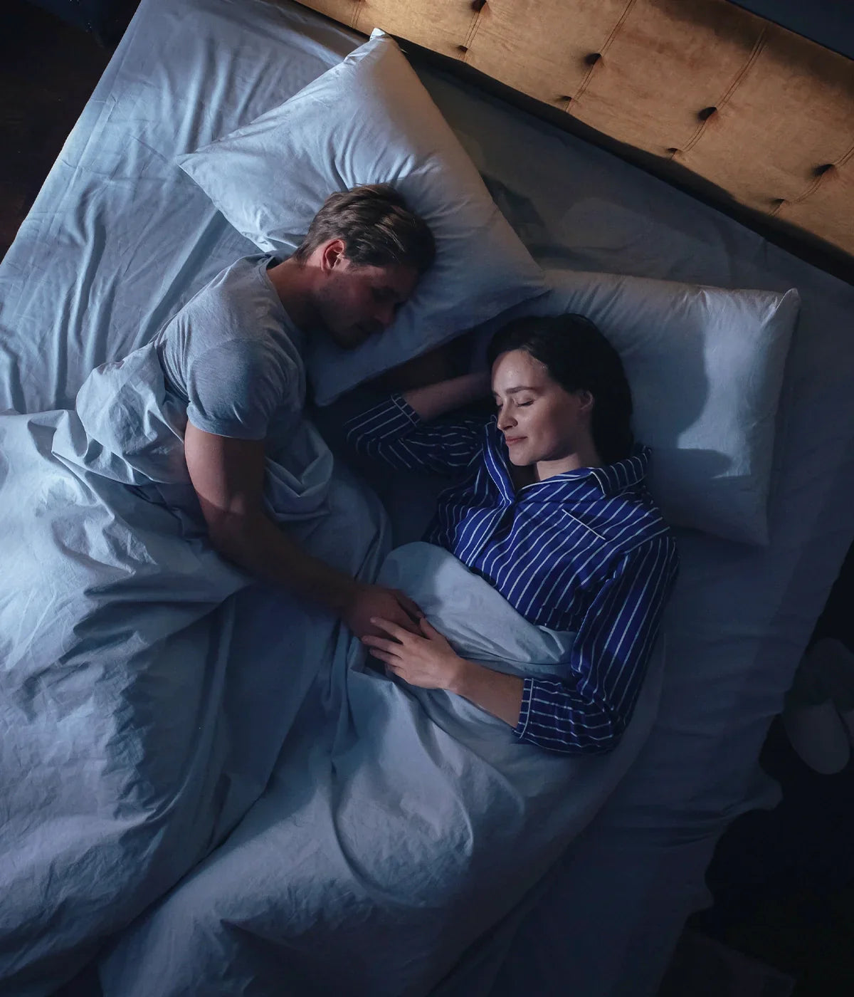 Husband wearing a grey t-shirt sleeping next to his wife wearing a dark blue striped pjs, holding hands in sleep while comfortably dozing on an Essentia Venti double sided organic latex mattress